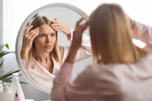 A woman looking at her forehead in the mirror and considering a brow lift in Miami